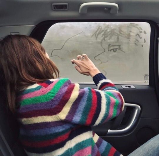 a woman sitting in the passenger seat of a car while drawing on the windshield window