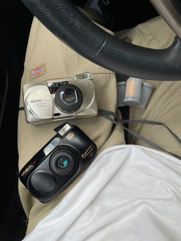 two cameras sitting on top of a car seat next to a steering wheel and cup holder