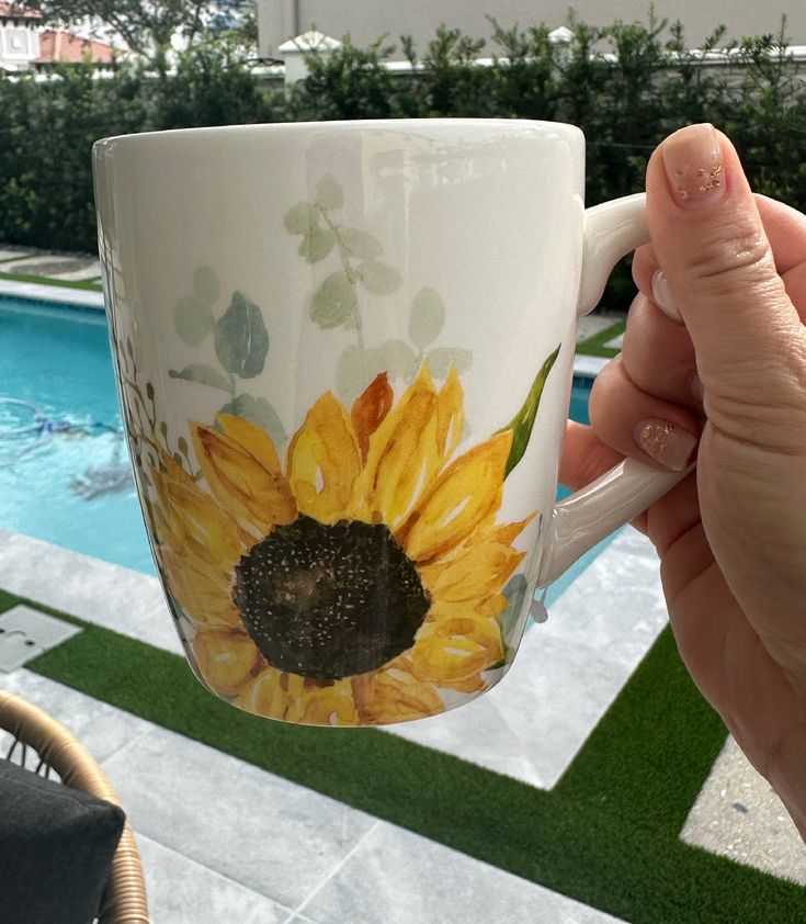 a hand holding a coffee cup with a sunflower painted on it next to a swimming pool