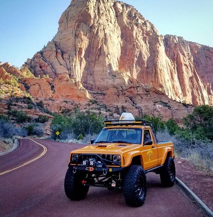 a yellow jeep driving down a road in front of a large rock formation with a light on it's roof