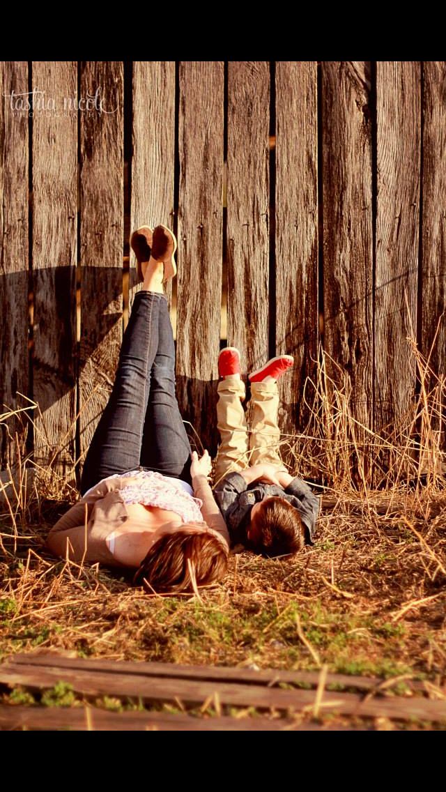 two people laying on the ground in front of a wooden fence with their feet up