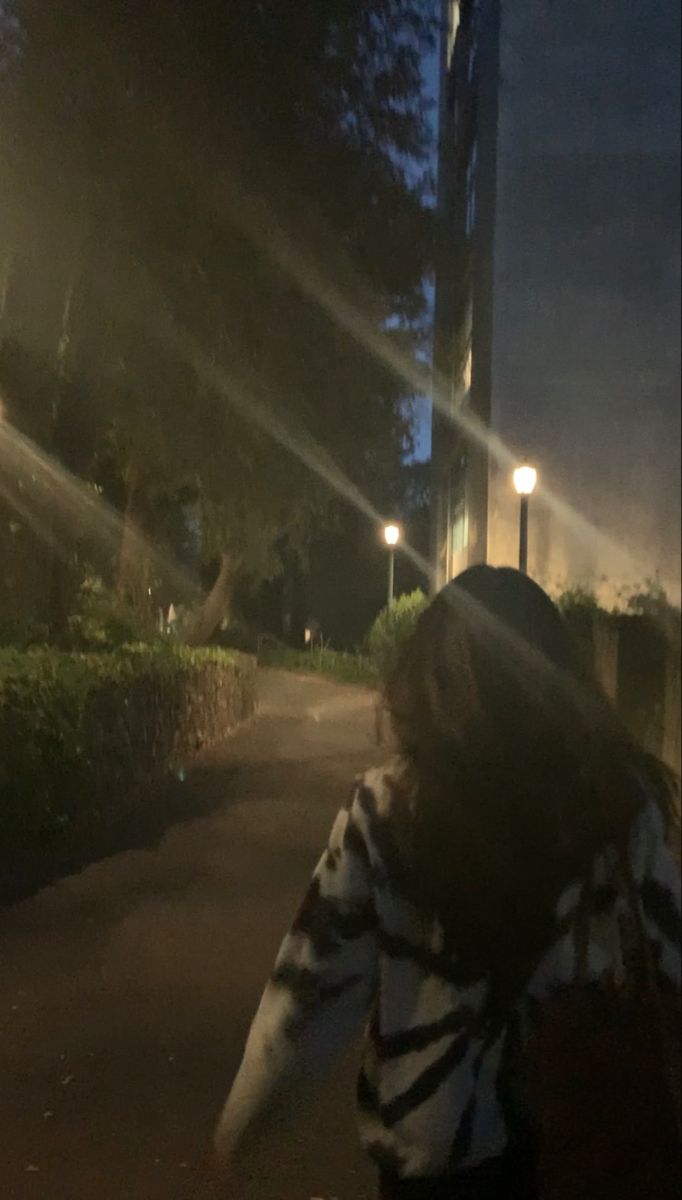 a woman walking down a street at night with her back to the camera and lights on