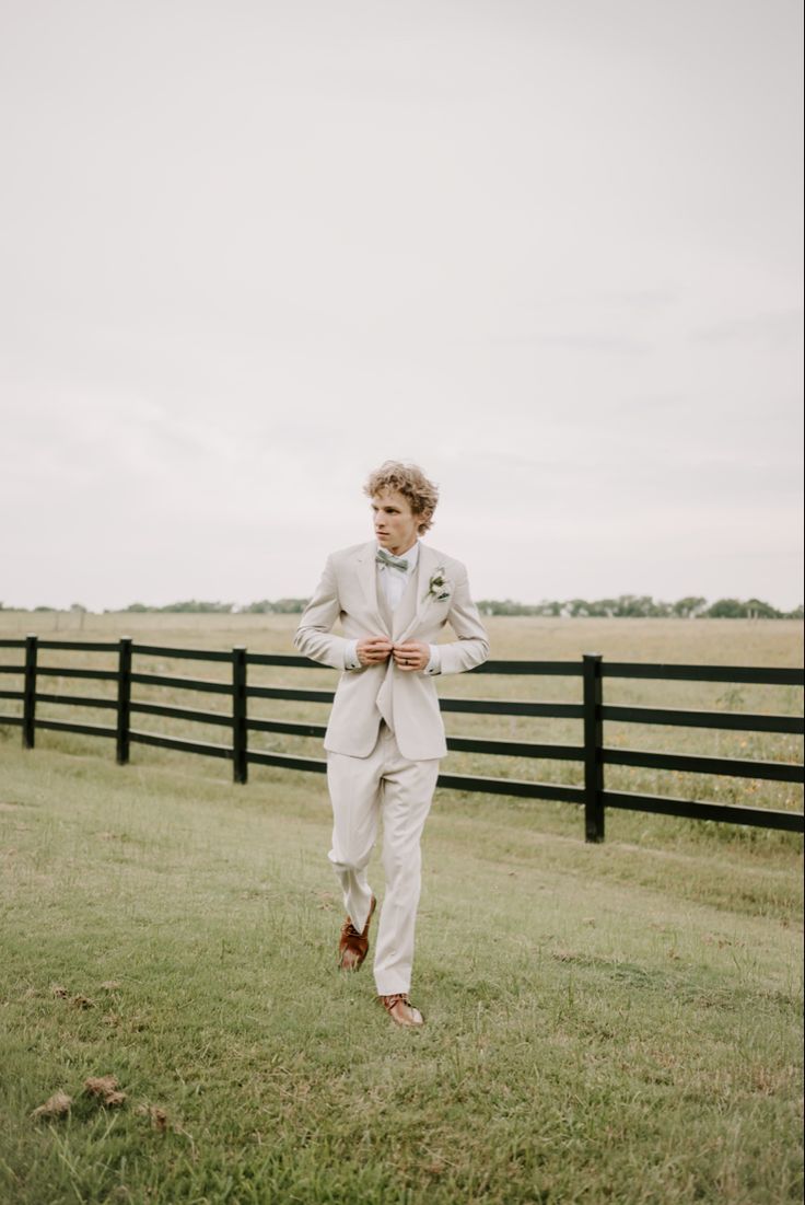 a young man in a white suit and tie standing on the grass near a fence