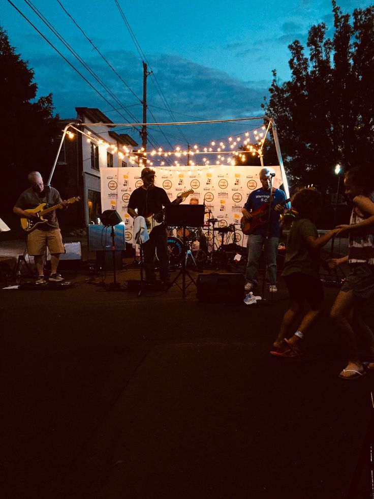 a group of people are playing music on the street at night with string lights strung over them