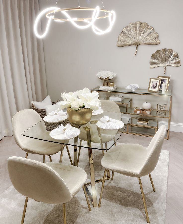 a glass dining table with white chairs and flowers on the top, surrounded by gold accents