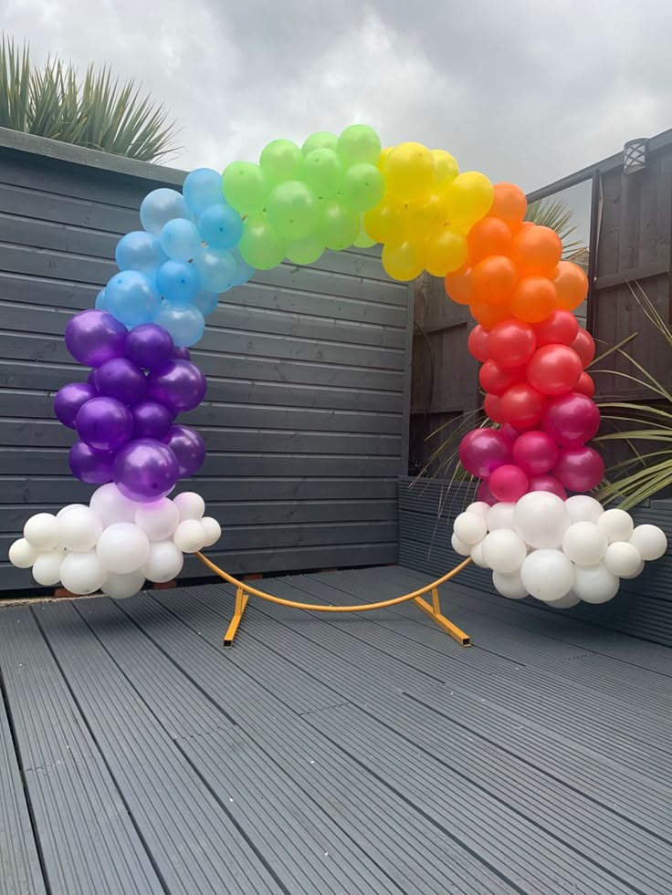 a rainbow arch made out of balloons on a deck with a potted plant in the background