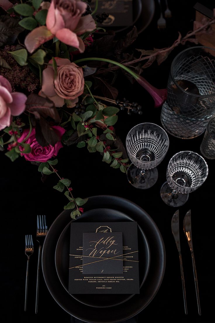 the table is set with black and gold plates, silverware, and pink flowers