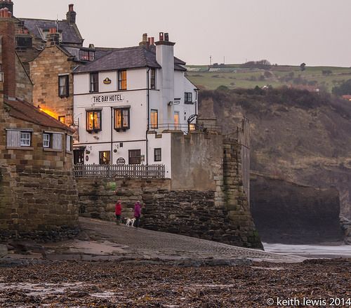 two people are walking on the beach next to some buildings and an old stone wall