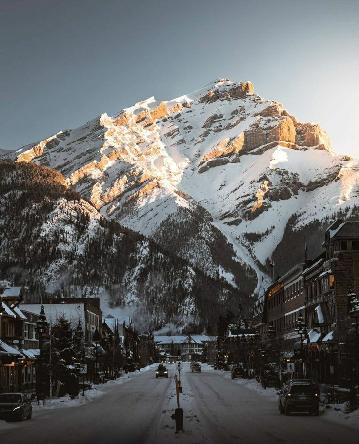 the sun shines on snow covered mountains and buildings along a city street in front of a snowy mountain