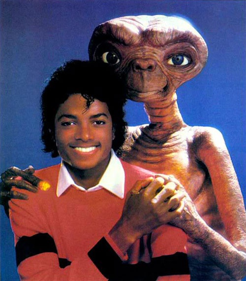 the man is posing with an alien in front of him and another creature behind him