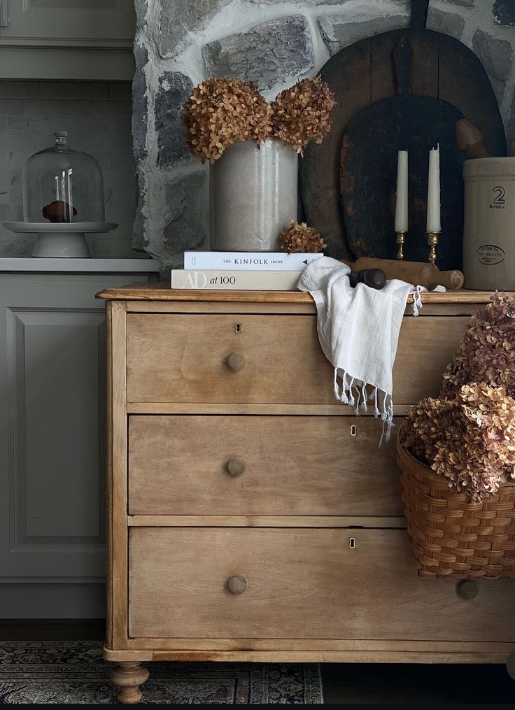 a wooden dresser topped with books next to a stone wall and vases filled with flowers