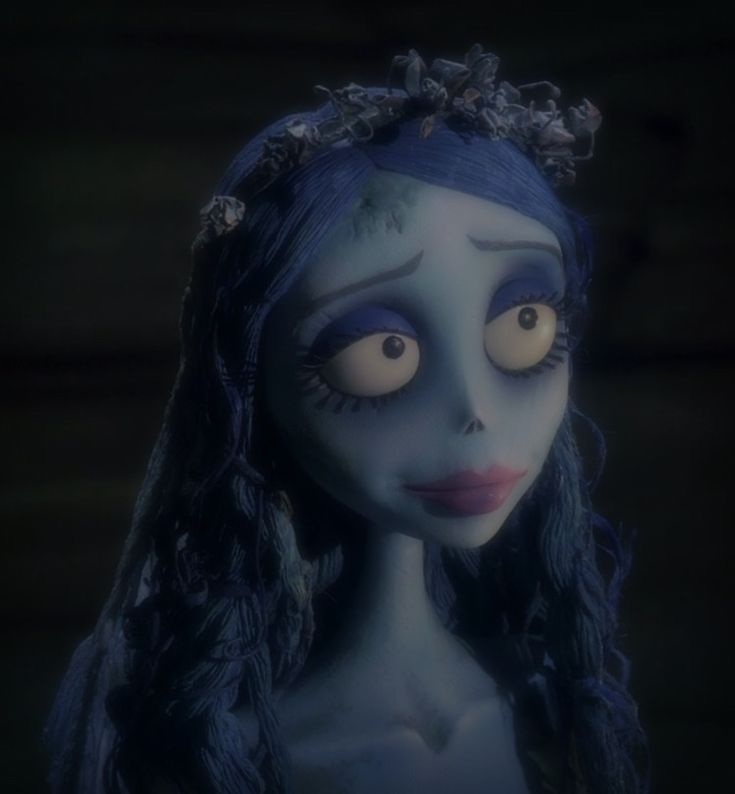 a creepy doll with blue hair and makeup looks at the camera while standing in front of a dark background