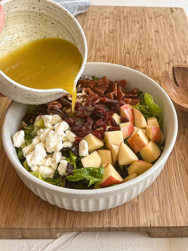 a person pouring dressing into a bowl filled with fruit and vegetables on a cutting board