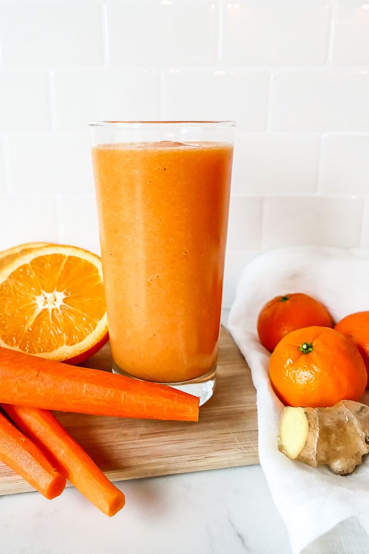 carrots, an orange and a glass of juice sit on a cutting board next to some cut up fruit