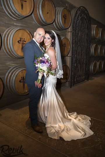 a bride and groom pose in front of barrels at their winery wedding venue, which is decorated with wine casks