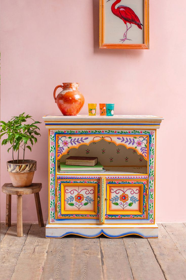 an old dresser painted with bright colors and birds on the top, next to a potted plant