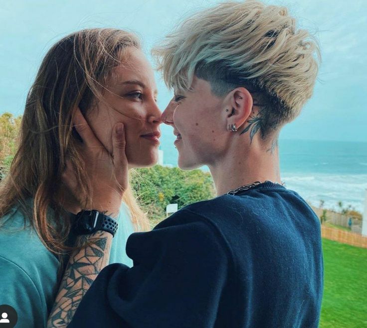 two young women are kissing each other in front of the ocean and grass, with one woman's face covered by her hands