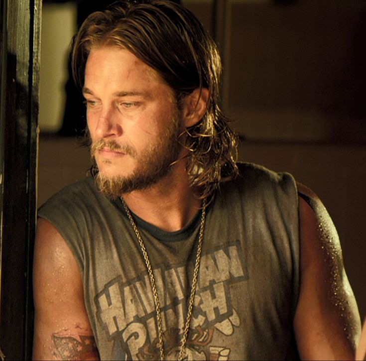 a man with long hair and beard wearing a tank top standing in front of a door