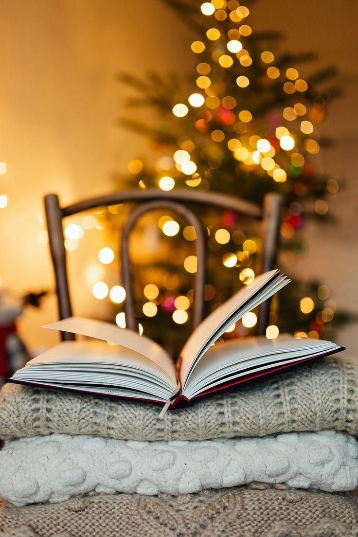 an open book sitting on top of a pile of blankets next to a christmas tree