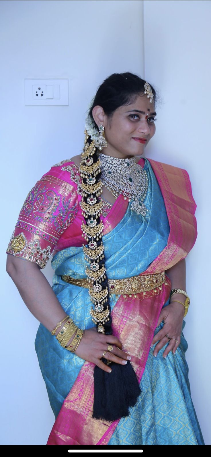 a woman in a blue and pink sari with gold jewelry on her neck, standing next to a white wall