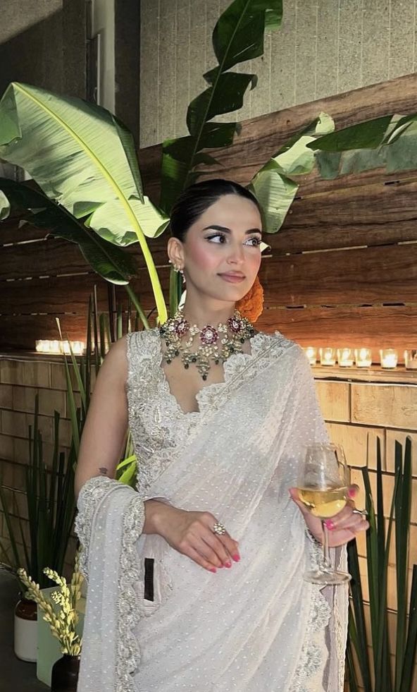 a woman wearing a white sari holding a glass of wine in front of plants