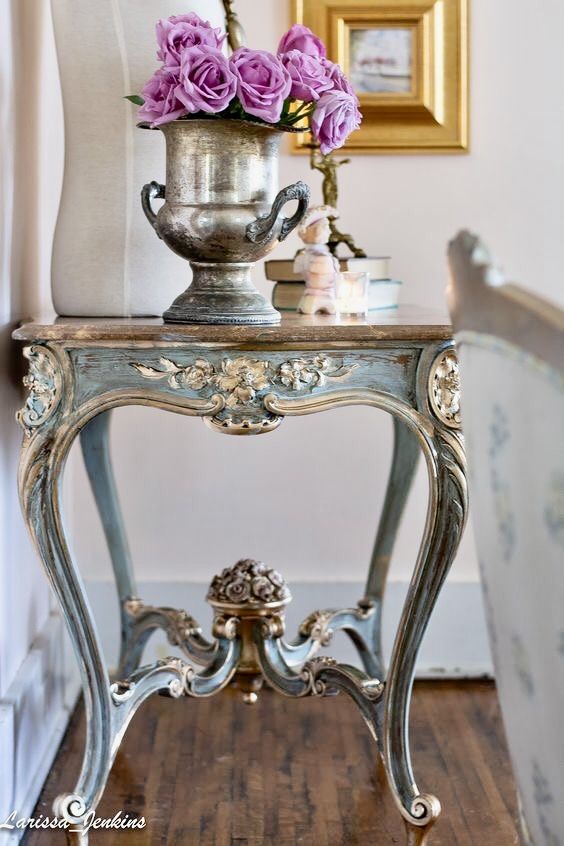 a silver vase with purple flowers on top of it sitting on a table next to a chair