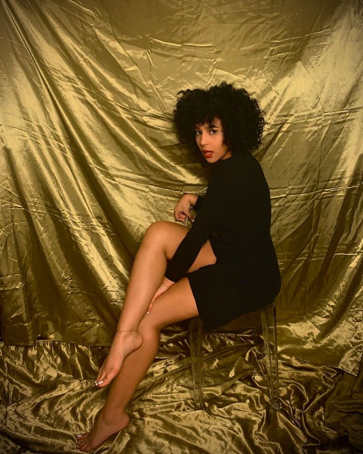 a woman is sitting on a chair in front of a gold sheet and posing for the camera