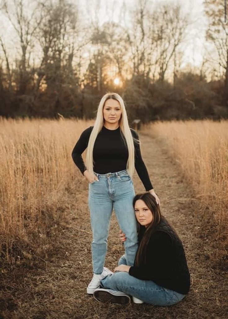 two women sitting on the ground in front of tall grass and trees, one with long blonde hair