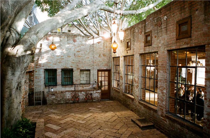 an empty courtyard with brick walls and trees growing over it, in front of a stone building