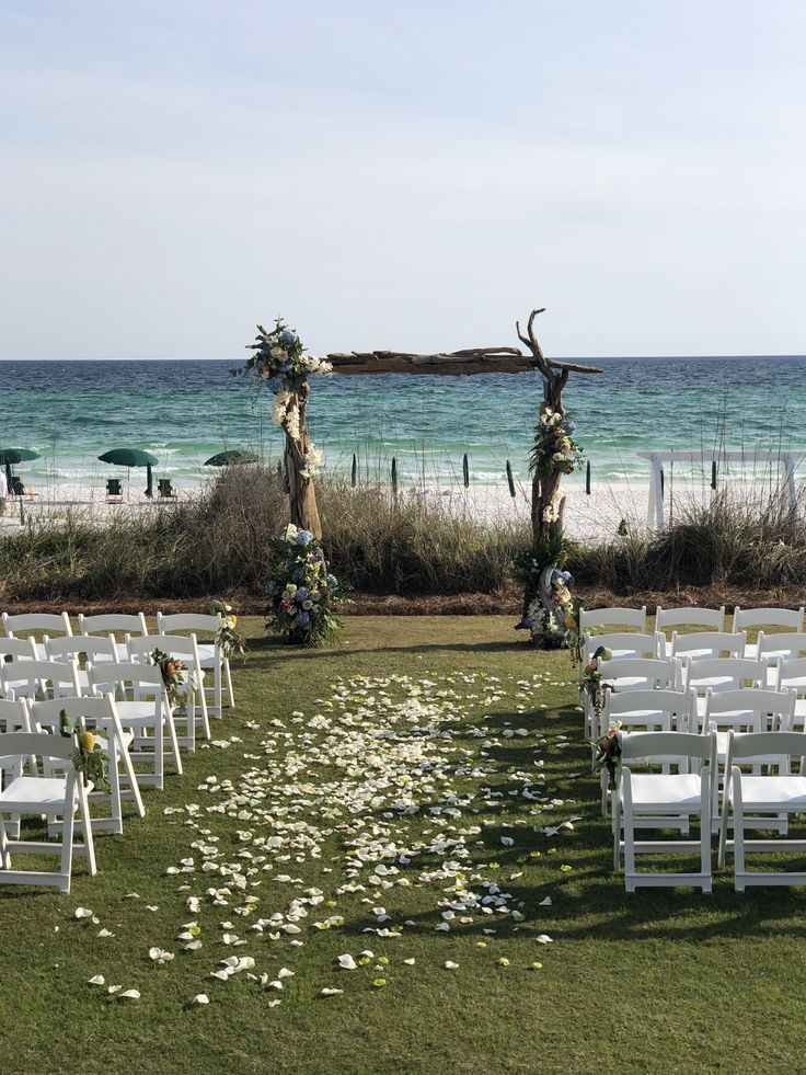 an outdoor ceremony set up with white chairs and flowers on the grass by the beach