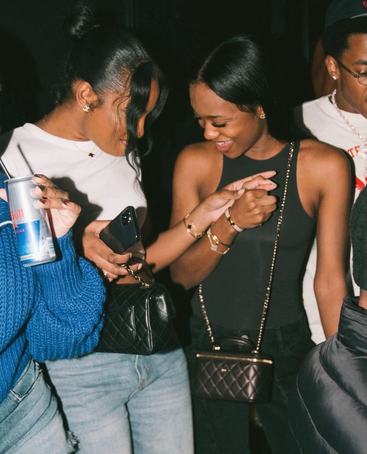 two women standing next to each other looking at their cell phones while one woman holds her purse