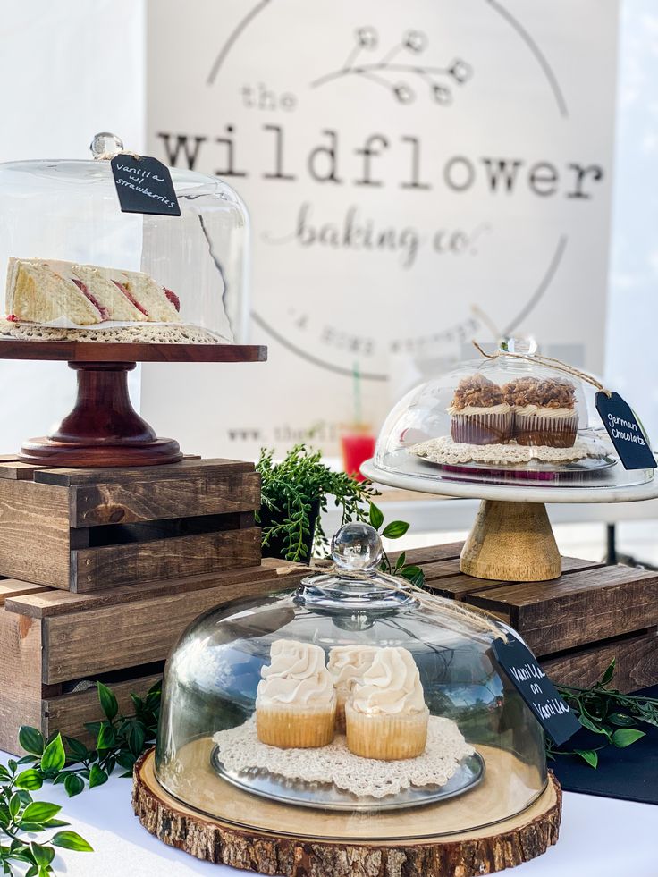 two cakes under a glass dome on top of wooden trays next to a sign that says wildflower baking co