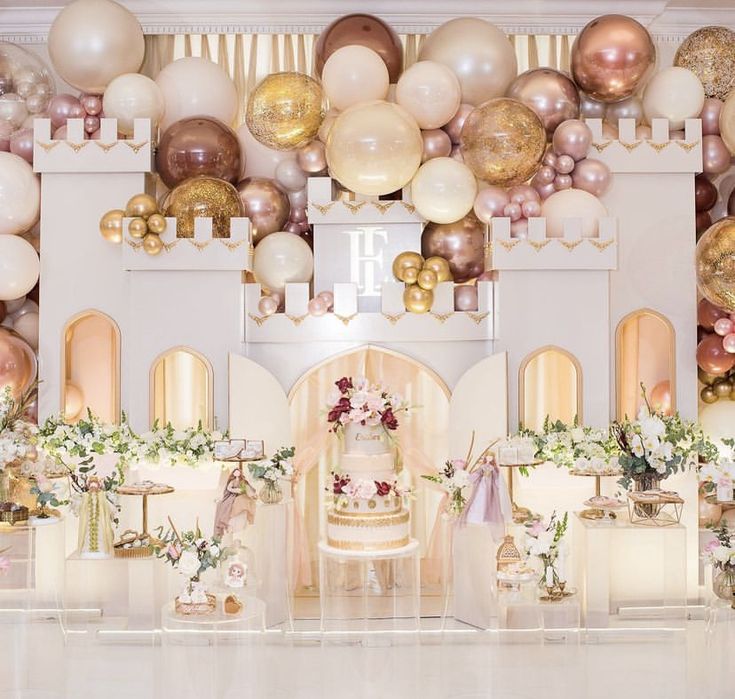 a table topped with cake and lots of balloons in front of a wall filled with gold, white and pink decorations