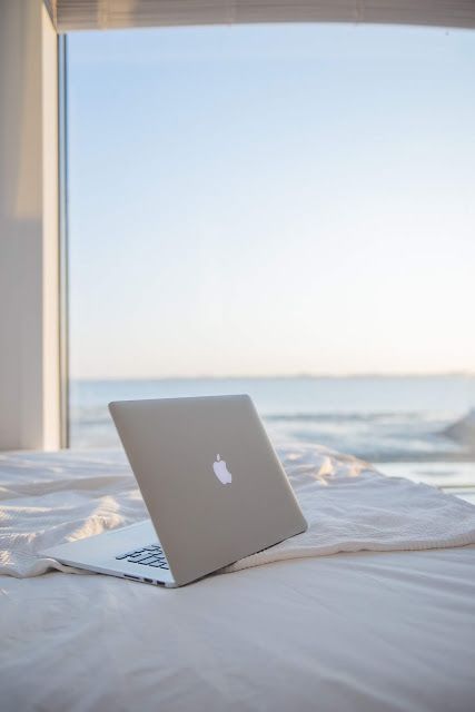 an open laptop computer sitting on top of a white bed next to a window overlooking the ocean