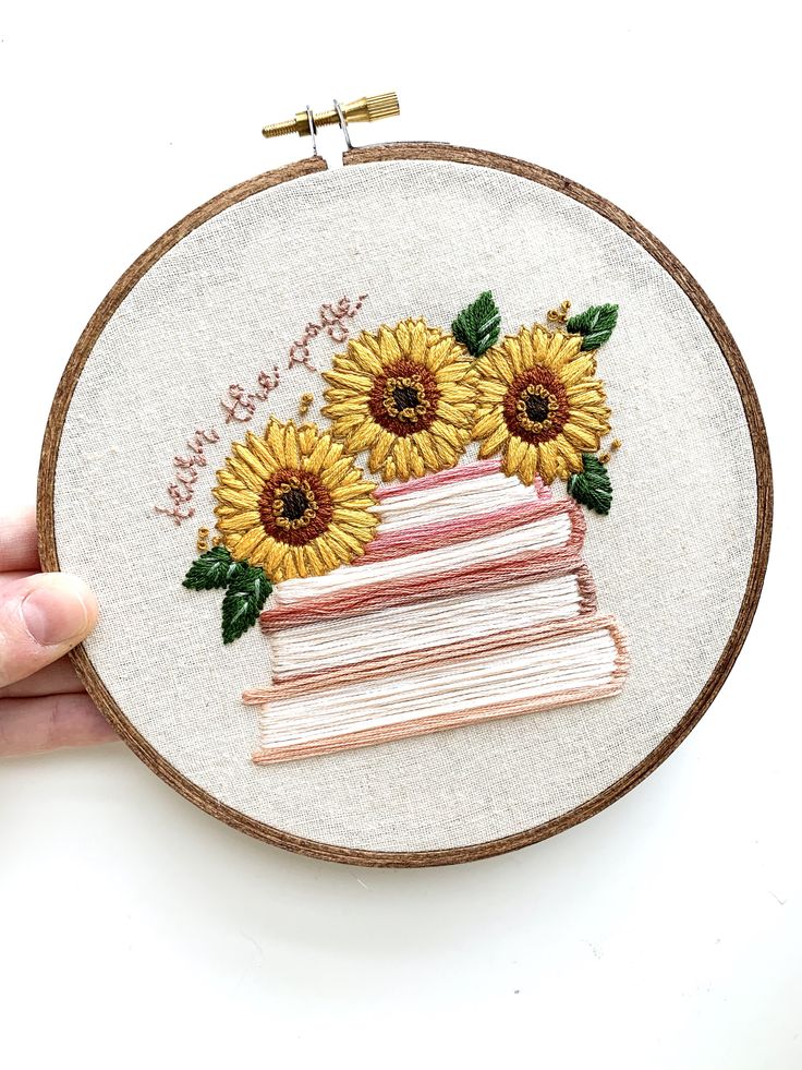 a hand is holding a cross - stitch hoop with a cake and sunflowers on it