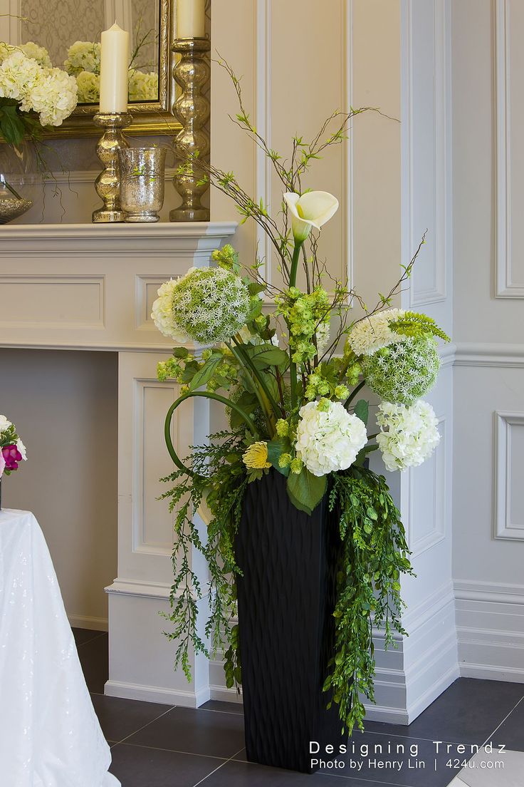 a black vase with white flowers and greenery on the table in front of a fireplace