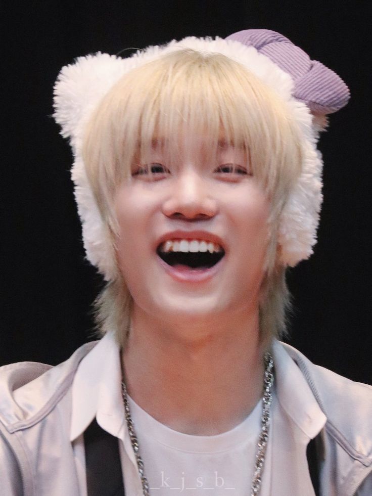 a young man with blonde hair wearing a furry hat and smiling at the camera while standing in front of a black background