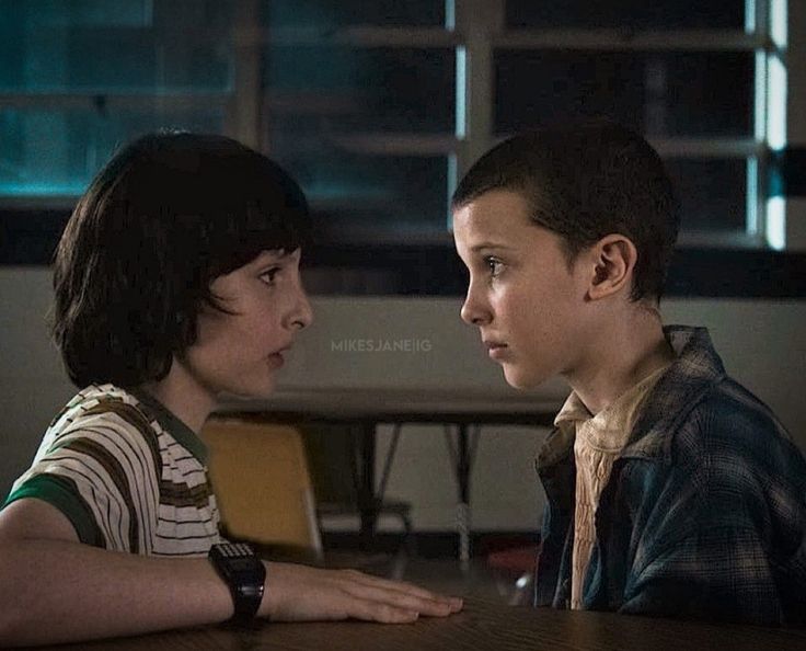 two young boys sitting at a table talking to each other