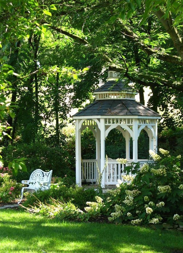 a white gazebo surrounded by lush green trees and shrubbery, with two chairs in the foreground