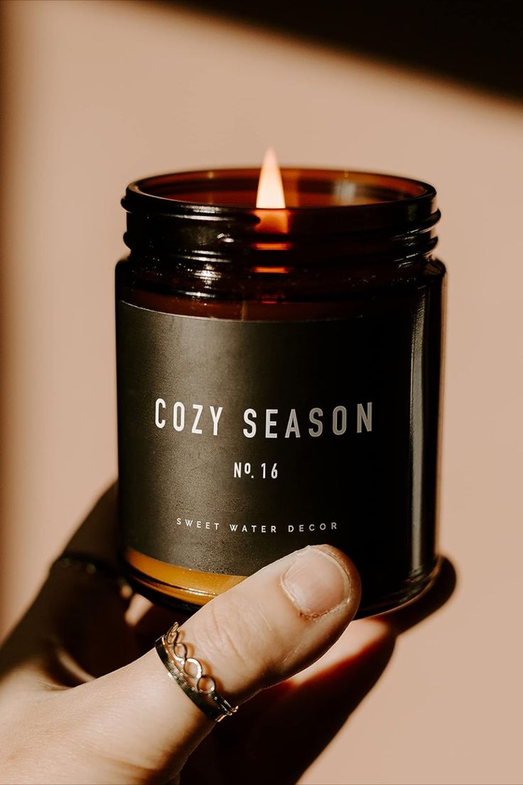 a hand holding a candle that says cozy season no 16 in front of a pink background