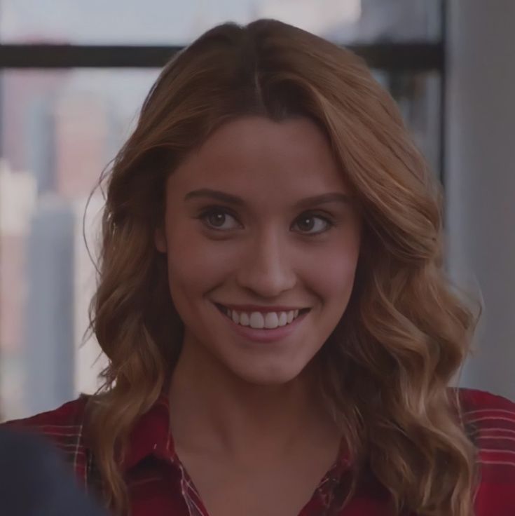 a woman with blonde hair smiling and wearing a red shirt in front of a window