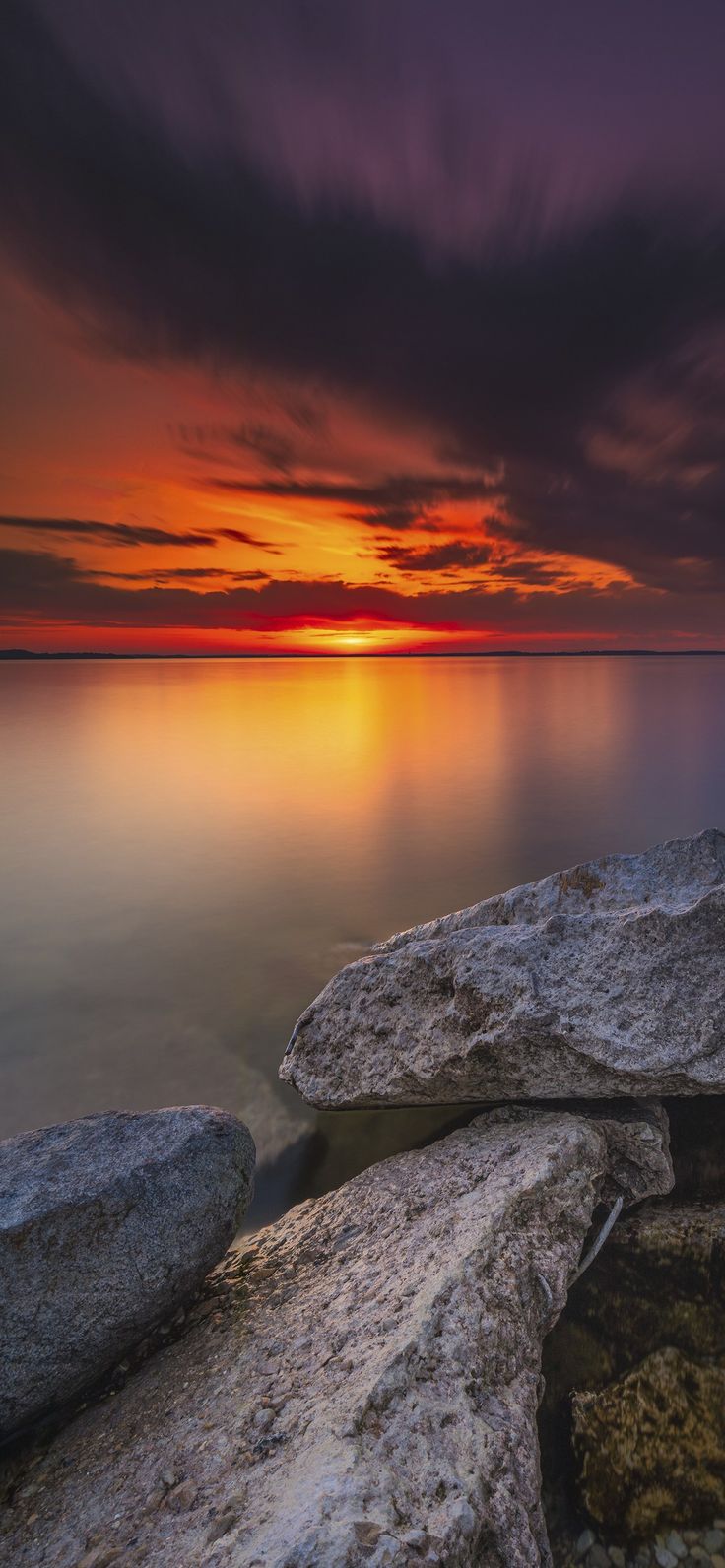 the sun is setting over the ocean with rocks in front of it and water below