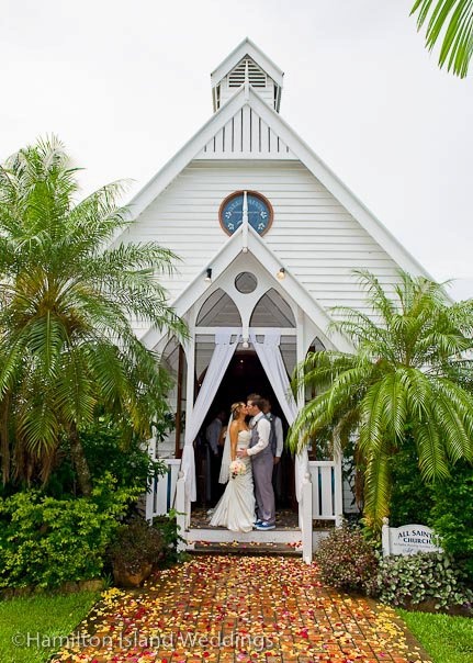 a bride and groom standing in front of a white church with fall leaves on the ground