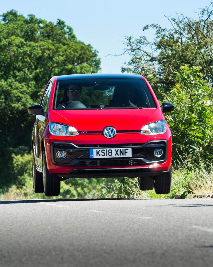 a red volkswagen car driving down the road with trees and bushes in the back ground