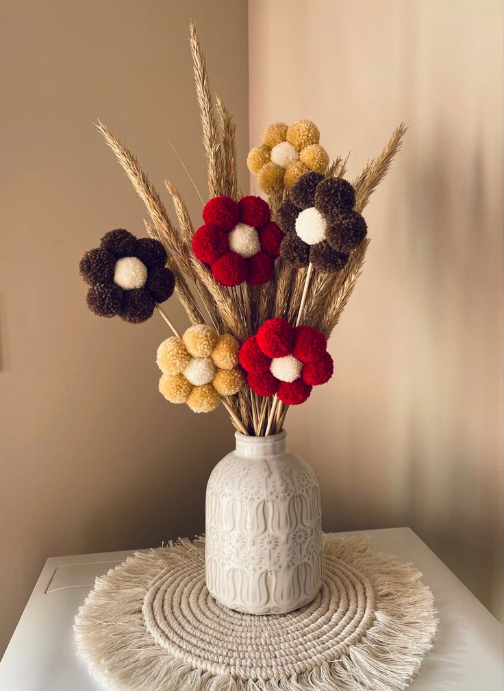 a white vase with red, yellow and brown flowers in it sitting on a table