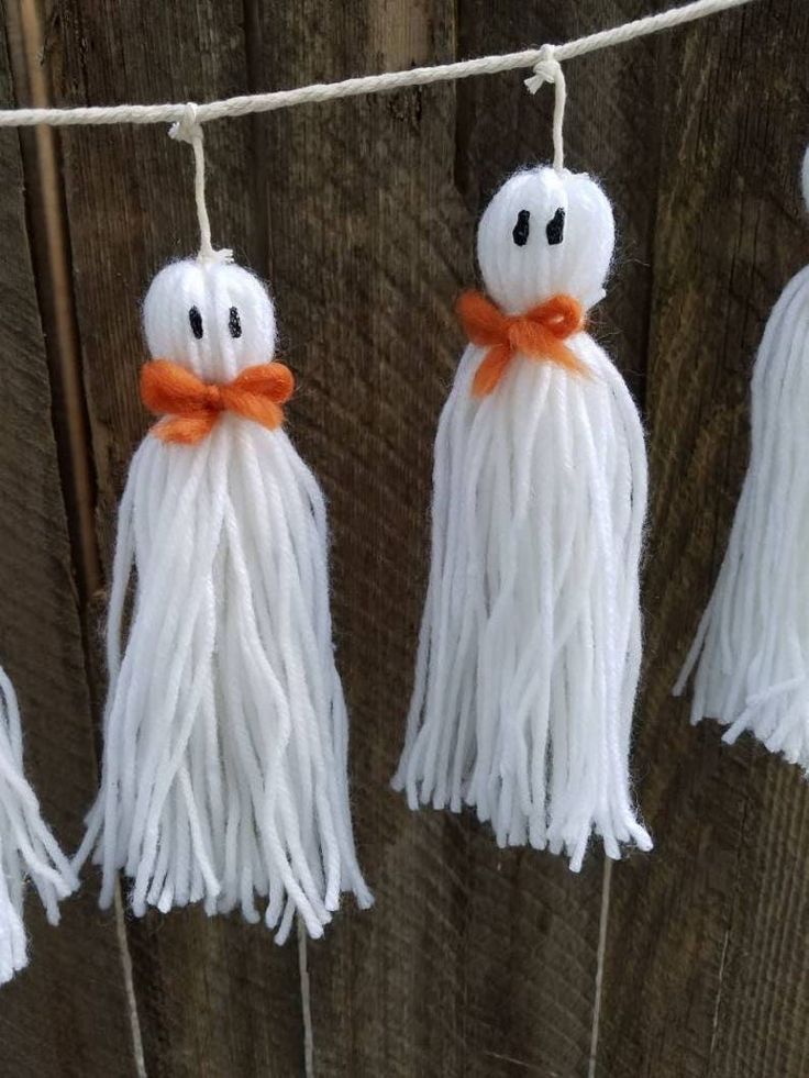 three white tasselled ghost decorations hanging from a line on a wooden fence with orange ribbon