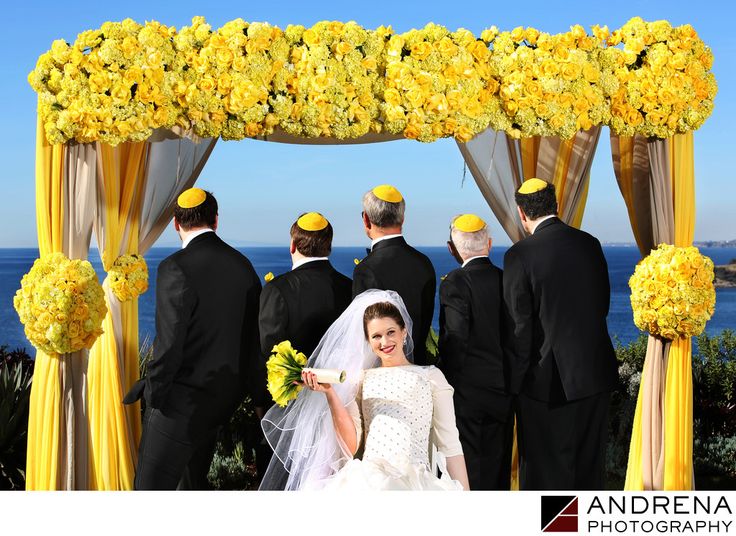 a bride and groom standing under a yellow flowered arch with their wedding party in the background