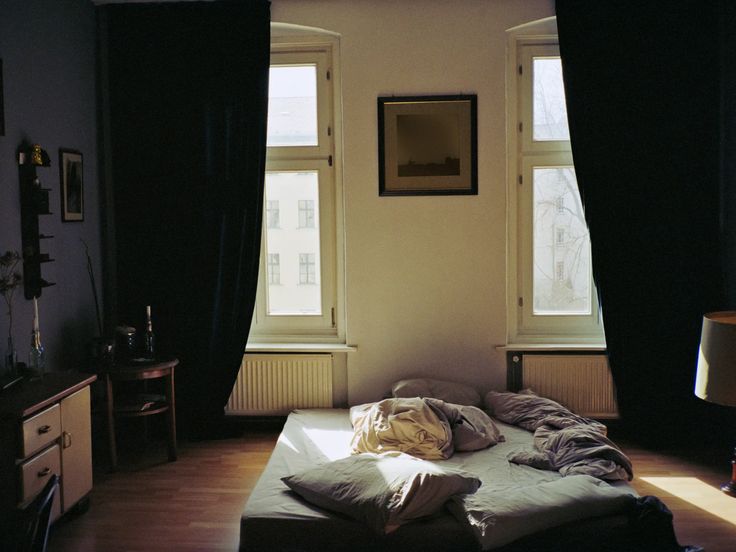 an unmade bed in a dark room next to two windows