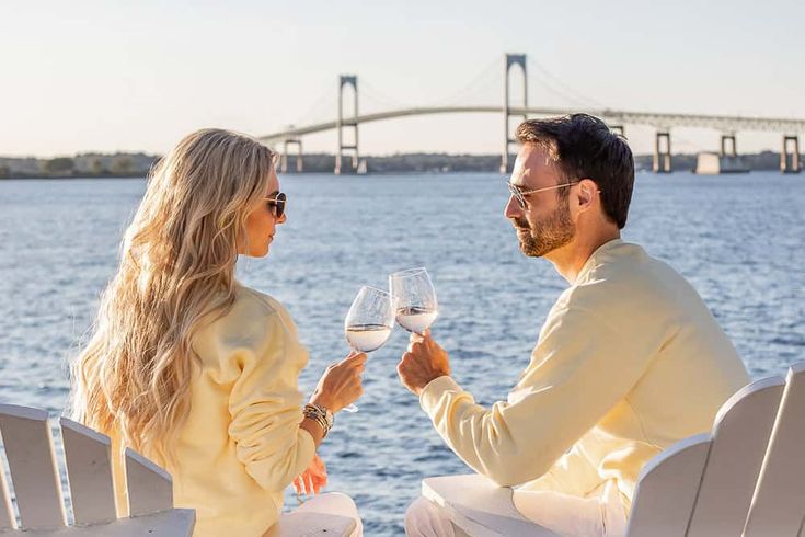 a man and woman sitting next to each other holding wine glasses in front of the water