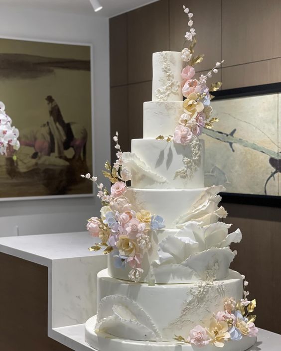 a white wedding cake with pink and blue flowers on top sits on a table in front of a painting